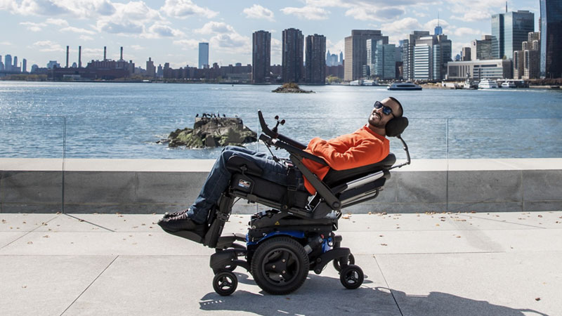 A man in a wheelchair leans back and smiles at the edge of a body of water surrounded by a city line