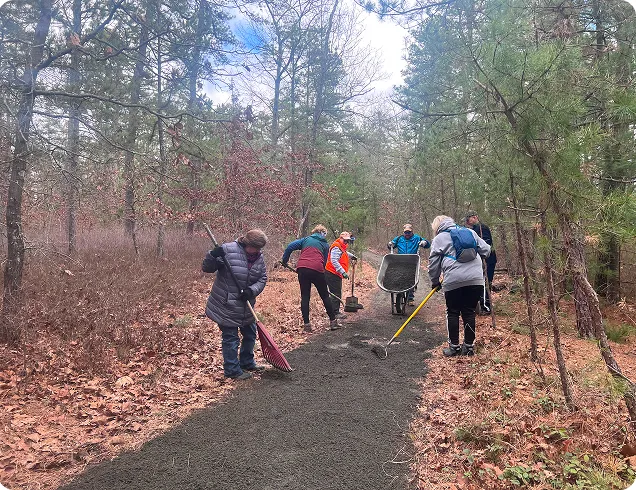 People help restore a pathway through the woods using rakes, shovels, and a wheelbarrow